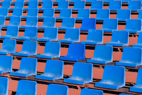 Empty blue plastic stadium seats are arranged in rows on a wooden bleacher structure, illuminated by bright sunlight in an outdoor setting