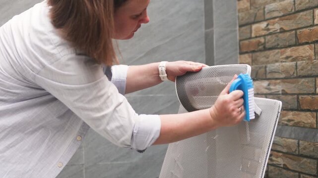 Cleaning with brush shows woman scrubbing grey office chair headrest with blue bristles. She uses circular motions for deep cleaning with brush to remove dust and stains from mesh fabric.