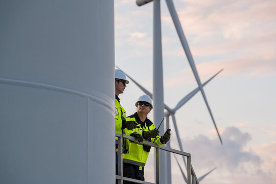 Diverse industrial technicians with tablet and walkie-talkie inspecting wind farm during evening shift, green energy concept.