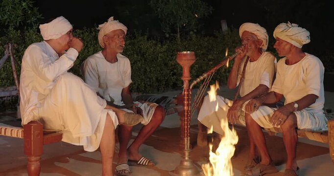 Indian old aged man wear white kurta pajama sitting on woven cot bed enjoy desi life at rural area. Happy three male group talk speak share gossip do hookah hold pipe spend night time village place
