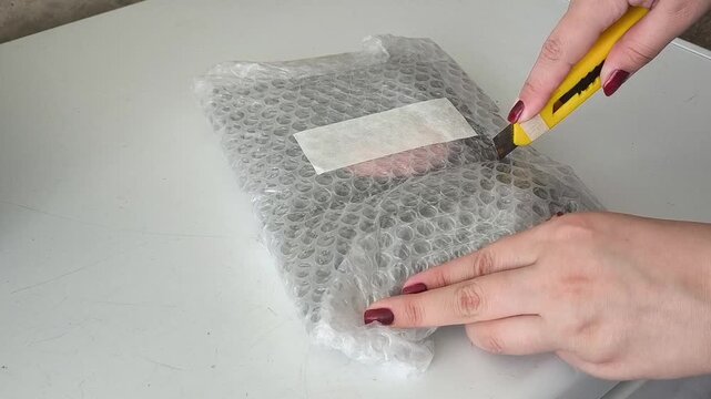 Close-up of hands unpacking transparent wrapped parcel with white stripe. Minimal delivery and ecommerce packaging scene on neutral gray surface.