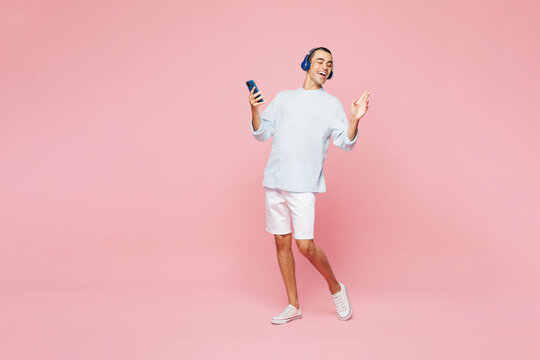 Full body young middle eastern man he wear casual blue cotton shirt listen to music in headphones use mobile cell phone isolated on plain pastel light pink wall background studio. Lifestyle concept