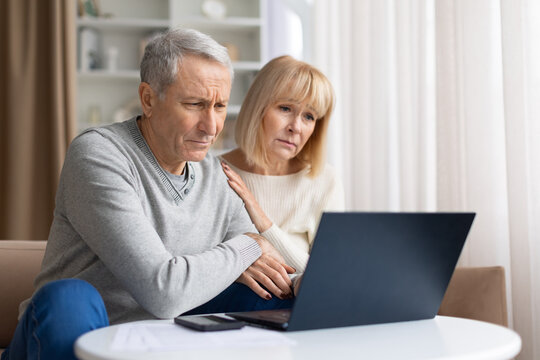 Concerned senior couple looking at laptop screen at home, reacting to bad online news or financial problem during retirement lifestyle indoors