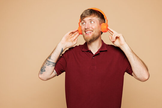 Young smiling stylish happy fun student man he wears red shirt casual clothes listen enjoy music in headphones isolated on plain pastel light beige wall background studio portrait. Lifestyle concept.