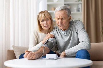 Fototapeta premium Concerned senior couple checking blood pressure monitor at home, worry elderly spouses focusing on healthcare, hypertension and elderly wellness indoors