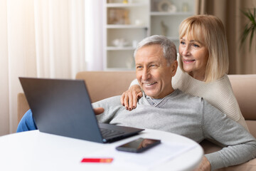 Fototapeta premium Relaxed mature couple watching laptop together on sofa at home, happy elderly spouses enjoying online entertainment and cozy retirement lifestyle indoors
