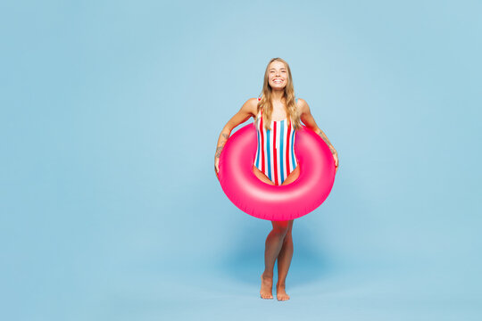 Full body young smiling satisfied woman she wears bikini swimsuit rest near hotel pool hold in hand pink rubber ring look camera isolated on plain blue background. Summer vacation sea sun tan concept.
