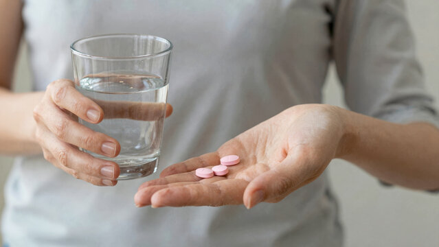 Close-up of a person holding a glass of water and several pink pills in their palm, ready to take medication
