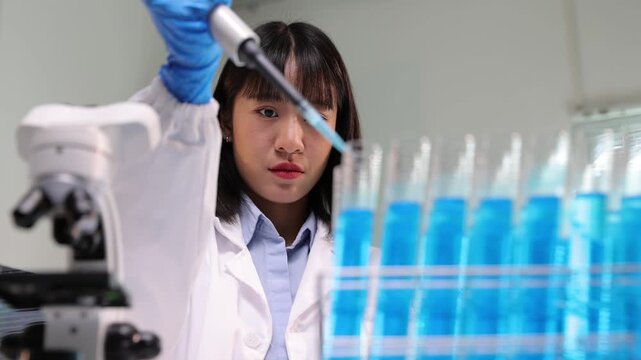 Young Asian female research scientist pipetting a blue liquid into test tubes. Laboratory equipment and microscope are in the background.