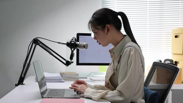 Asian female podcaster live streaming from her home office. Young Asian woman wearing an apron typing on a laptop and talking into a professional microphone, managing her online small business.