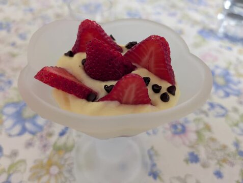 Dessert cremoso servito in coppa di vetro, guarnito con fragole fresche e gocce di cioccolato su tovaglia floreale in stile casalingo