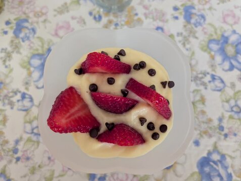 Dessert cremoso in ciotolina bianca, decorato con fragole fresche e gocce di cioccolato su tovaglia floreale dai toni pastello