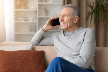 Fototapeta premium Happy senior man talking on smartphone while relaxing on sofa at home, enjoying pleasant conversation in cozy domestic living room environment