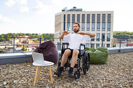 A man with a beard sits in a wheelchair on a rooftop terrace, performing arm exercises with a laptop nearby and beanbags scattered around
