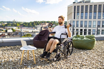 Obraz premium A man with a beard in a wheelchair is exercising with dumbbells on a rooftop terrace with a city view