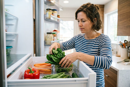 Woman organizing fresh green vegetables inside the bottom drawer of an open refrigerator