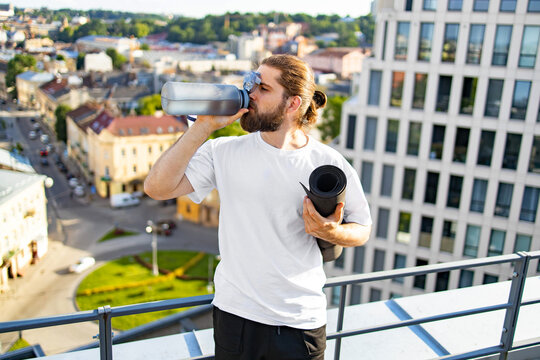 A bearded man with his hair in a bun drinks from a large water bottle while holding a rolled-up yoga mat on a rooftop overlooking a city