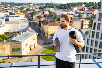 A man with a beard and long hair tied back stands on a rooftop holding a yoga mat and a water bottle, overlooking a city © sofiko14
