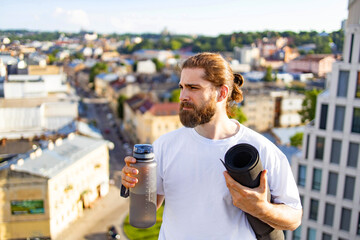 A man stands on a rooftop, holding a water bottle and yoga mat while looking out over the city skyline © sofiko14