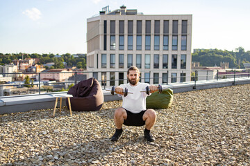 Obraz premium A bearded man with long hair performs dumbbell squats on a rooftop terrace with a city view and beanbag chairs