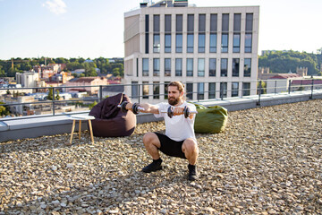 Obraz premium A man with a beard and tied-back hair performs dumbbell squats on a gravel-covered rooftop with a city view and modern building in the background