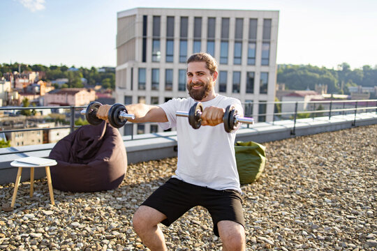 A smiling man with a beard and bun is performing bicep curls with dumbbells on a rooftop terrace with a city view