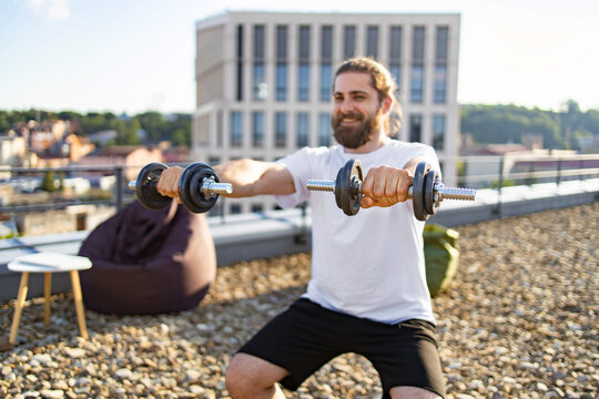 A smiling man with a beard and long hair is performing bicep curls with dumbbells on a rooftop terrace with a city view