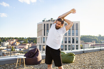 Obraz premium A man with a beard and bun hairstyle performs a shoulder stretch on a rooftop terrace with a city view and modern building in the background