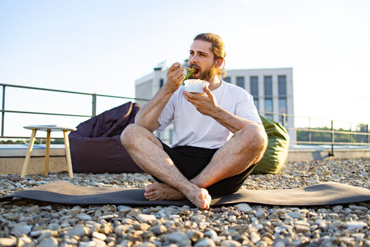 A man with a beard and tied-back hair sits cross-legged on a yoga mat on a gravel rooftop, eating a salad from a plastic container
