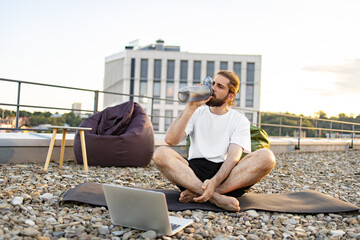 Obraz premium A man with a beard and long hair sits cross-legged on a yoga mat on a gravel rooftop, drinking from a water bottle while a laptop is open in front of him