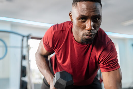African American man doing bent-over row gripping hexagonal dumbbell wearing red tee in gym