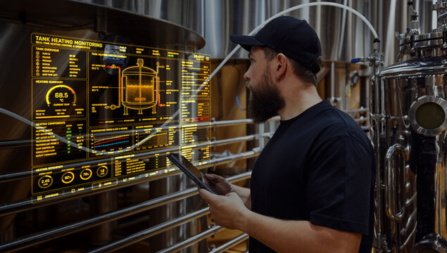 Brewery technician checking tank heating data on digital monitoring panel near production equipment. Concept of thermal control, smart brewing, energy monitoring and process automation.