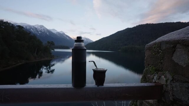 A thermos and mate cup rest on a railing with a serene Patagonian lake and mountain backdrop. Ideal for travel promotions or cultural pieces.