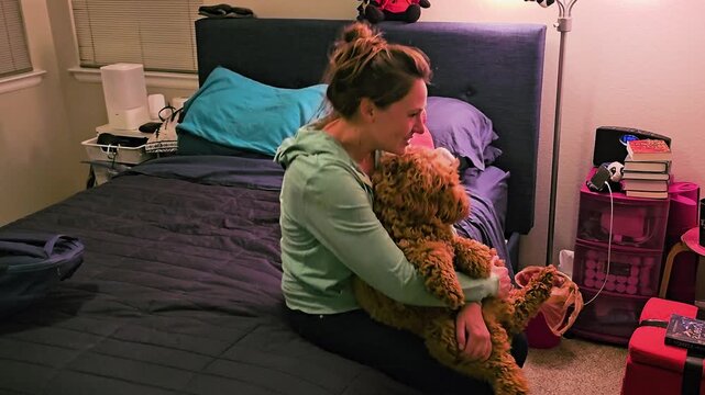 Woman cuddling and dancing with her goldendoodle on a bed in a warm bedroom, playful pet owner bonding moment, curly dog, cozy home lifestyle, love, and companionship.