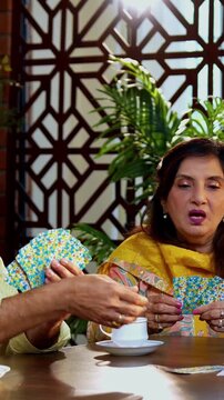 Indian senior adults playing cards game while seated at dining table in cozy modern home, laughing and bonding joyfully, Asian old aged friends celebrating cheerful indoor reunion with fun activity