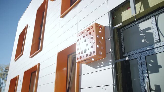 Modern architectural detail features vibrant orange accents on wall. Construction site view captures facade cladding process under sun