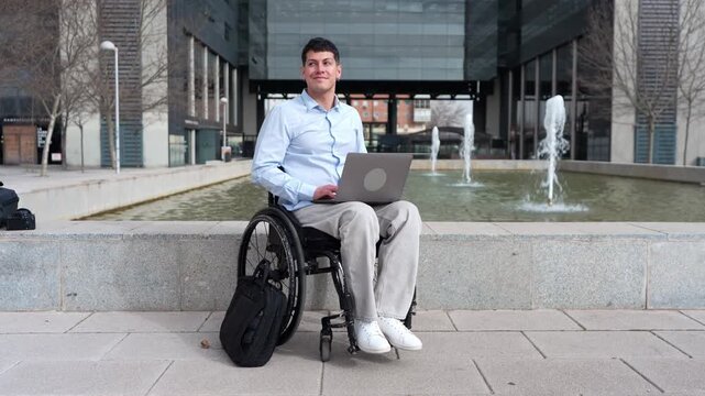 Young disabled businessman in a wheelchair working remotely on a laptop. Paralyzed male entrepreneur teleworking in a modern city plaza