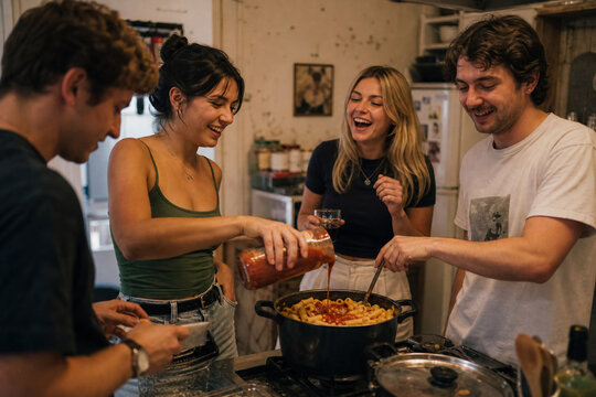 Four young adults cooking pasta and laughing together in a home kitchen, suitable for content about friendship, home cooking, and happy social gatherings.
