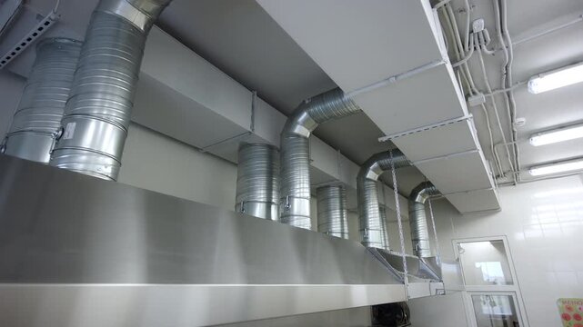 Industrial ventilation system features metal pipes and exhaust hoods. Complex ductwork stretches across ceiling of commercial kitchen