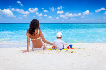 Naklejka premium Rear view of a mother and her little baby son playing together in the sand of a beautiful beach with white sand and turquoise ocean