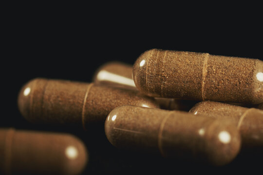 Macro still life of transparent capsules with brown vitex fruit powder on a black textured background. Selective focus on capsules with bright reflections. Pure herbal supplement for female wellness.