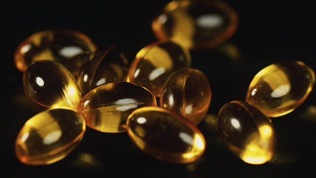 Macro still life of Vitamin D3 capsules in a heap on a black textured background. Selective focus on golden glowing capsules with bright reflections. Essential micronutrient for winter wellness.