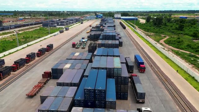 Wide top-down drone shot of a large container depot with rows of stacked shipping containers and trucks moving along a rail freight corridor. Modern logistics infrastructure supporting cargo transport