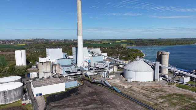 A back tracking drone shot of the massive chimney at Studstrup Power Station, overlooking green fields and the Kaloevig Bay Area