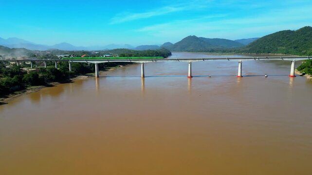 Cinematic aerial drone footage of the green Lao-China high-speed train traveling across a bridge over the Nam Song River near Vang Vieng, Laos. Scenic landscape with lush greenery and mountains as mod