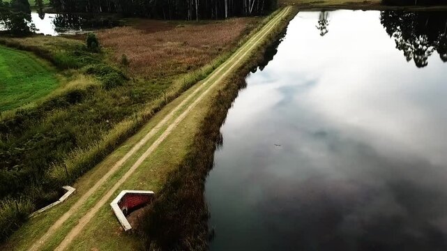 Aerial view of a drone sweeping across a lake with a pathway for vehicles in a field with green grass