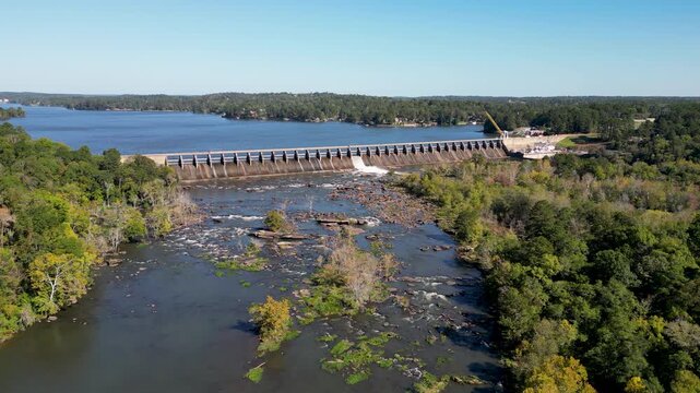 Oliver Dam on Chattahoochee River with spillway releasing water into rocky downstream channel and Lake Oliver reservoir above hydroelectric facility operated by Georgia Power smooth clockwise drone or