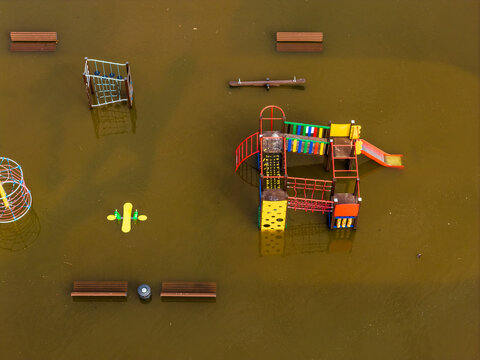 Aerial view of a flooded children's playground with colorful climbing frames and wooden benches submerged in murky brown water in Kedainiai, Kaunas County, Lithuania.