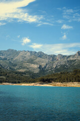 Obraz premium Blue lake water reflecting a bright sky with rugged mountains and a green forest on a sunny day