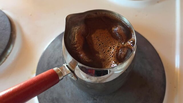 Coffee brewing in a metal pot on a stove. Foam bubbling as the drink reaches a boil. Traditional process of making hot dark caffeinated beverage for breakfast.